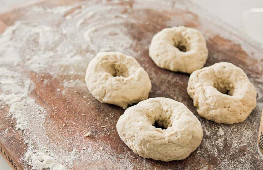 Four raw bagels on a floured wooden surface, ready for baking.